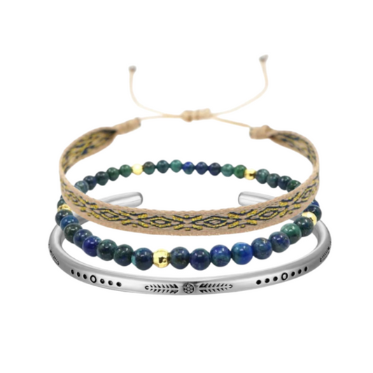 A wrist wearing three stacked bracelets: one beige woven with navy and gold pattern, one with green-blue beads and gold accents, and one engraved silver-toned metal cuff.