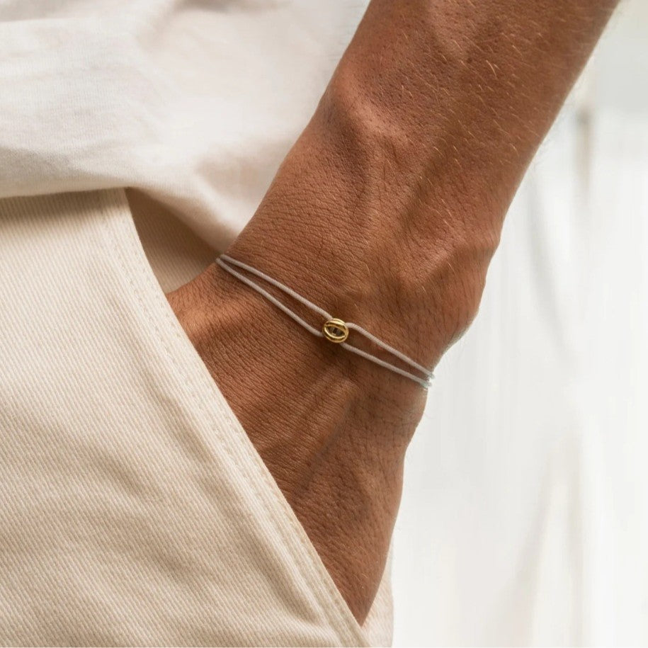 A person’s wrist adorned with a grey bracelet featuring a golden lucky charm, adding a subtle touch of elegance and positivity to their outfit.